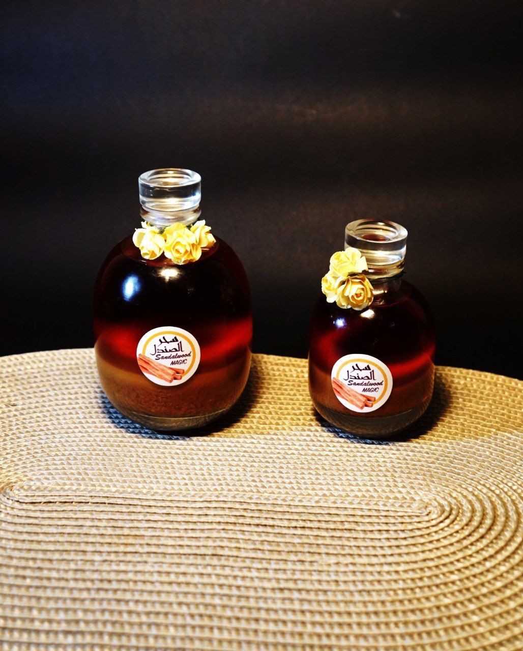 Two glass bottles labeled 'Sandalwood Magic' with yellow flower decorations on a woven mat.